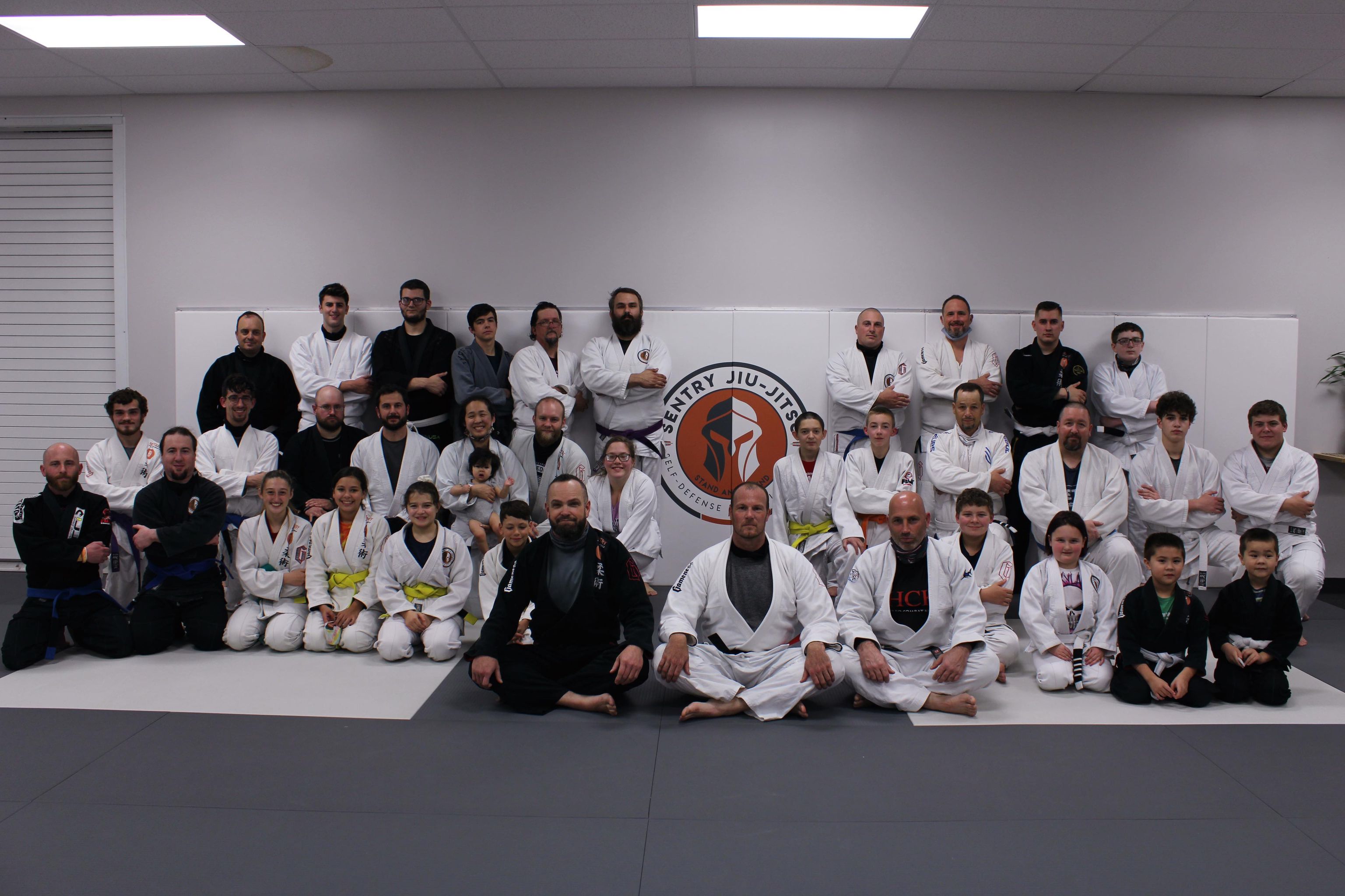 The Sentry Jiu-Jitsu team of kids and adults together on the mats in front of the academy logo